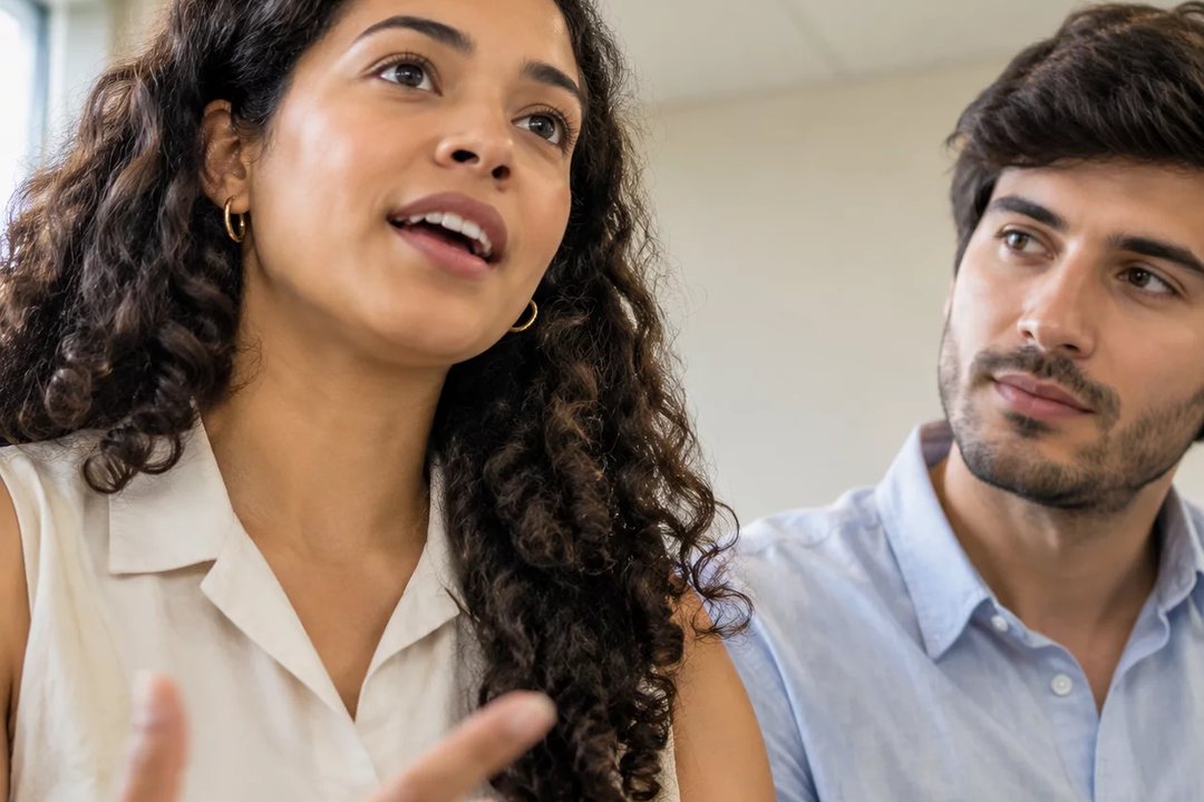 Três participantes conversam ao redor de uma mesa comprida numa sala clara de janelas altas